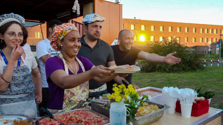 Pressebild: Menschen mit bunten Kochmützen geben an einem Stand im Abendlicht Essen aus
