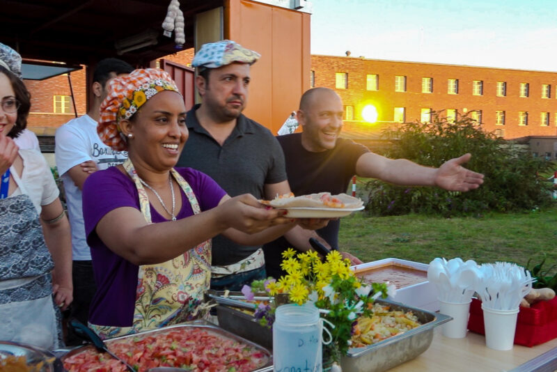 Pressebild: Menschen mit bunten Kochmützen geben an einem Stand im Abendlicht Essen aus